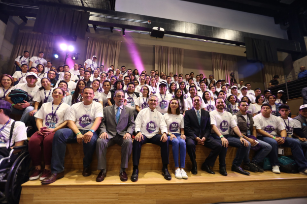 Gruppe von Menschen auf einer Bühne mit einer Person im Rollstuhl links, Vorhänge und Scheinwerfer im Hintergrund.