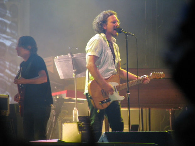 Ein Mann in einem weißen Hemd spielt Gitarre und singt in ein Mikrofon auf der Bühne, neben ihm ein anderer Mann in einem schwarzen Hemd, der Gitarre spielt; ein Piano, Kabel, Lautsprecher und Scheinwerfer sind im Hintergrund zu sehen.