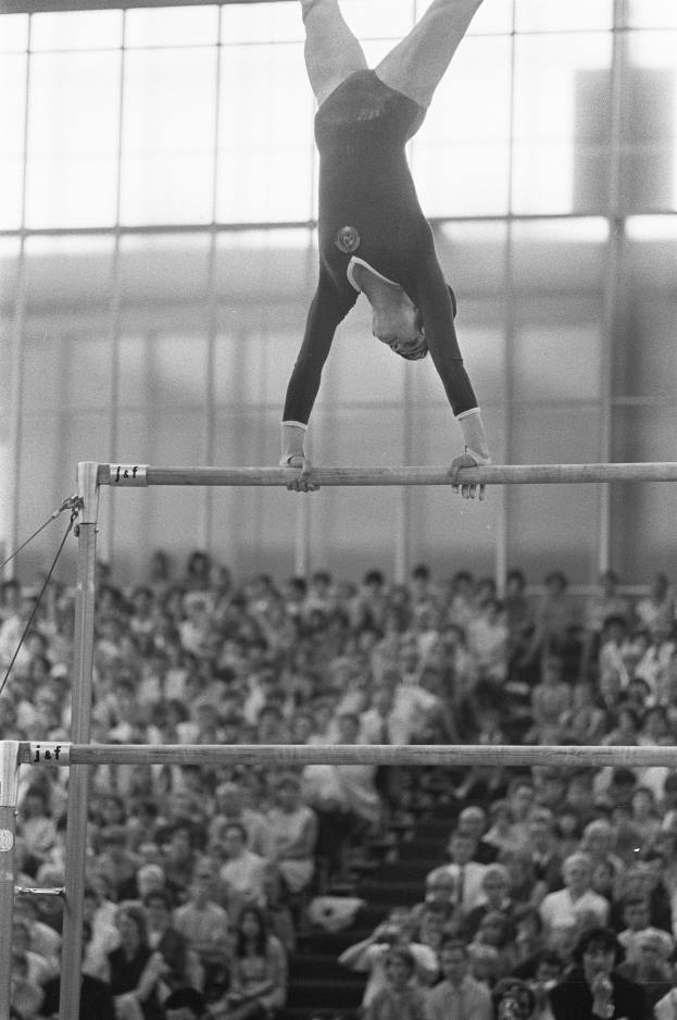 Eine Frau führt Gymnastik an den parallelen Stangen aus, vor einem sitzenden Publikum dargestellt in Schwarz-Weiß.