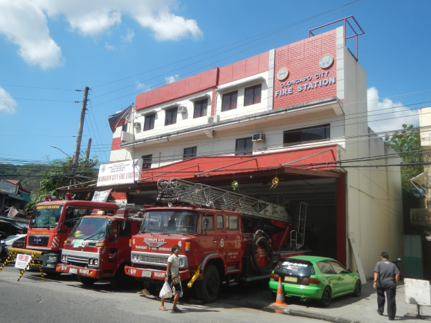 Gruppe von Feuerwehrautos vor einem Geb├Ąude mit Fenstern und Text, umgeben von Fahrzeugen, Verkehrskegeln, Passanten, Strommasten, B├Ąumen und einem bew├Âlktem Himmel.