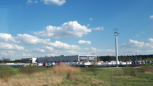 Eine Mercedes-Benz-Fabrik in Deutschland mit Fahrzeugen auf der Straße, Grünflächen, Gebäuden und einem Logo-Mast unter einem bewölkten Himmel.