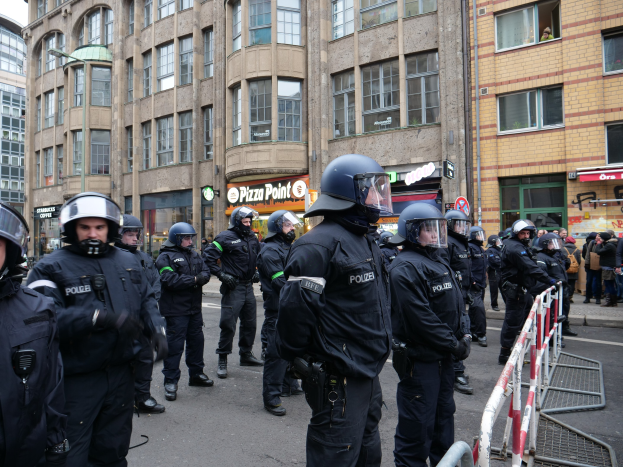 Gruppe von Polizeibeamten vor einer Menschenmenge hinter Absperrungen auf einer Straße mit Gebäuden, Schildern, Laternenpfählen und einem klaren blauen Himmel im Hintergrund.
