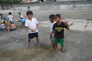 Eine Gruppe junger Jungs, die in einer Parkanlage stehen, einige halten Flaschen, mit Häusern, Bäumen und Pfählen im Hintergrund bei klarem blauem Himmel.