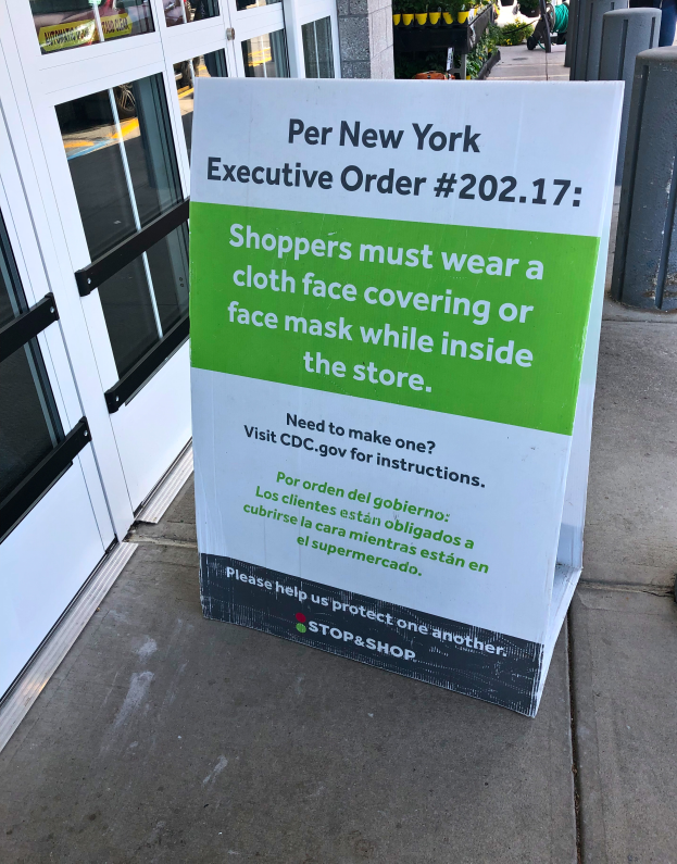 Ein Schild auf einem Bürgersteig vor einem Geschäft trägt die Aufschrift "Per New York Executive Order 202.17: Kunden müssen im Geschäft einen Stoffgesichtsschutz oder eine Gesichtsmaske tragen", mit einem Gebäude auf der linken Seite und Menschen, Pflanzen und anderen Gegenständen auf der rechten Seite.