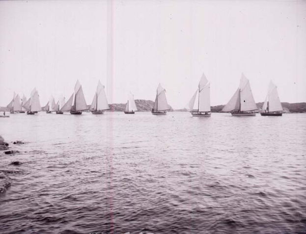 Schwarzes und weißes Foto von Segelbooten auf dem Wasser mit Hügeln im Hintergrund und einem klaren Himmel, als das New York Yacht Club bezeichnet.