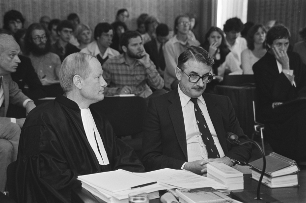 Richter des Obersten Gerichtshofs John Roberts in einer schwarzen Robe und ein anderer Mann in einem Anzug sitzen an einem Tisch mit Büchern, Papieren, einem Glas und einem Mikrofon und schauen auf eine Menge mit einer Wand und einem Vorhang im Hintergrund.