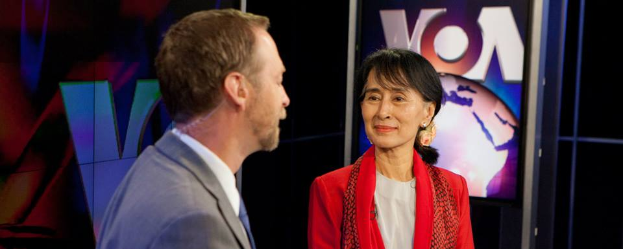 Ein Mann in einem grauen Blazer und weißem Hemd steht neben einer Frau in einem roten und weißen Kleid vor einem Fernseher, der den Text 'Aung San Suu Kyi auf VOA' anzeigt, vor einem schwarzen Hintergrund.