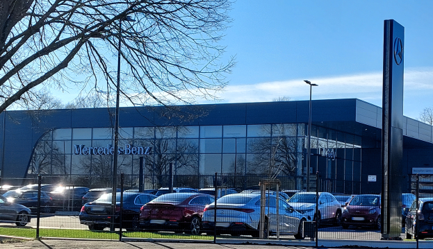 Ein Mercedes-Händler mit parkenden Autos vor einem Metallzaun, Gras, einem Schild, Straßenlaternen, einer Gruppe von Bäumen und einem bewölkten Himmel.