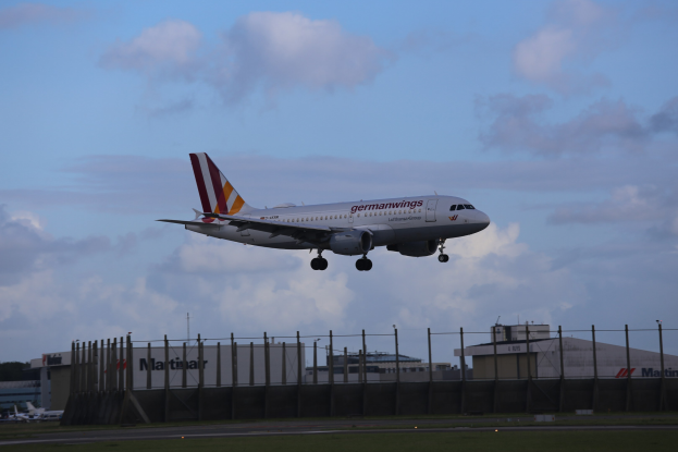Germanwings Airbus A320-200 beim Start vom Frankfurt Airport mit Gras darunter, einem Zaun im Vordergrund, Gebäuden und Bäumen im Hintergrund und einer bewölkten Himmel überkopf.