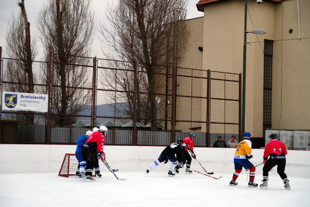 Menschen beim Eisschnelllauf auf einer Eisfläche mit Gebäuden, Bäumen, einer Straßenlaterne, einem Namensschild und Zäunen im Hintergrund unter einem Himmel.