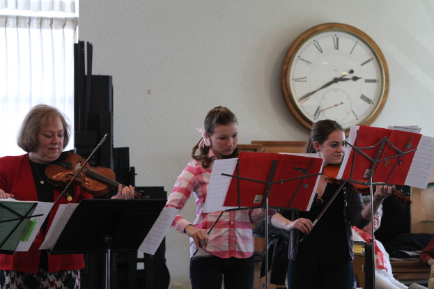 Drei Frauen, die auf der Bühne Geigen spielen, während eine Gruppe von sitzenden Frauen im Vordergrund zuhört; eine Uhr hängt an der weißen Wand im Hintergrund, mit einem weißen Vorhang in der linken Ecke.