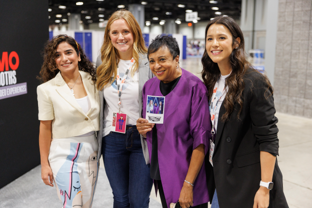 Vier Frauen auf ID-Karten lächeln gemeinsam auf einem Konferenzstand, mit einem Textschild links und Lichtern im Hintergrund.