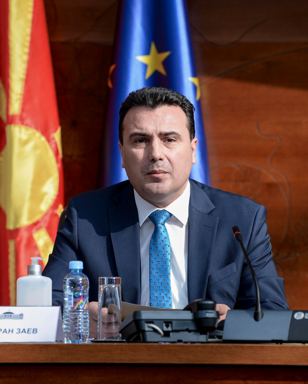 Ein Mann in Anzug und Krawatte sitzt an einem Tisch mit einem Mikrofon, Flaschen, einem Glas und einer Namensschilder, mit Flaggen im Hintergrund.