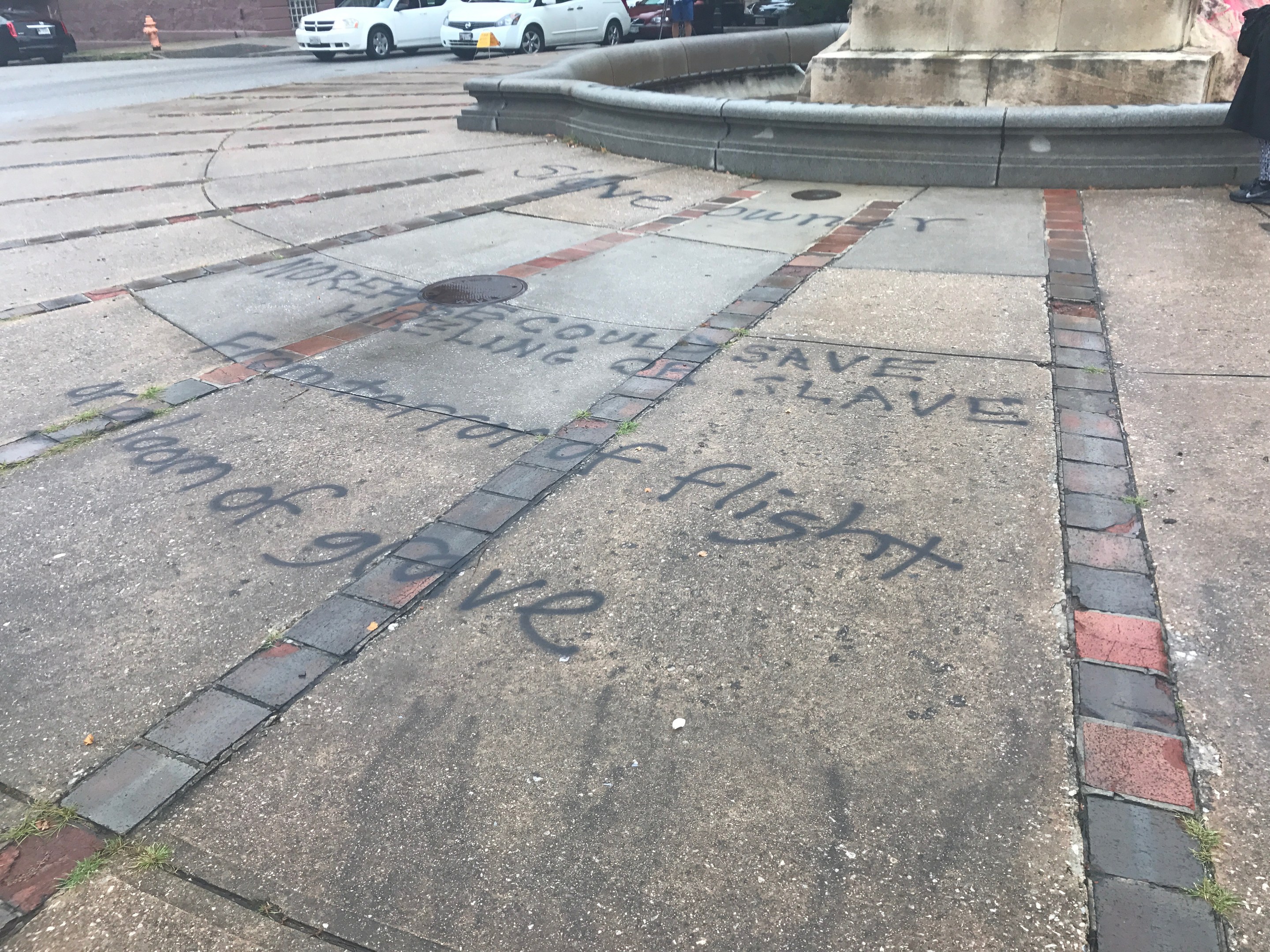 Geweg mit "Black Lives Matter" Graffiti in großen Buchstaben vor einem Gebäude, mit Fahrzeugen, Menschen, Bäumen, Pfählen und einer Wand in der Nähe.