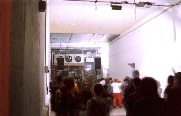 Leute tanzen in einem Raum mit Lautsprechern, einem Rohr und einer weißen Wand, unter Lichtern, einem Projektor und einem Eisenrahmen, mit verschiedenen Gegenständen im Hintergrund.