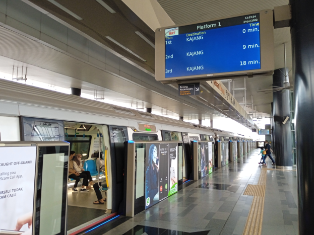 Eine U-Bahn fährt in einen Bahnhof ein, mit Fahrgästen im Inneren und anderen auf dem Bahnsteig, unter einer Decke mit Lampen und einem digitalen Display.