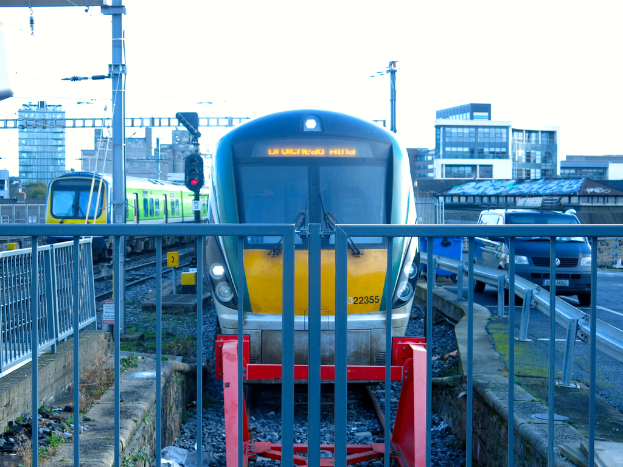 Zwei Züge auf Schienen mit einem Zaun im Vordergrund, Pfähle mit einem, der ein Signallicht hat, ein Auto auf der Straße rechts, Gebäude im Hintergrund und Himmel darüber.