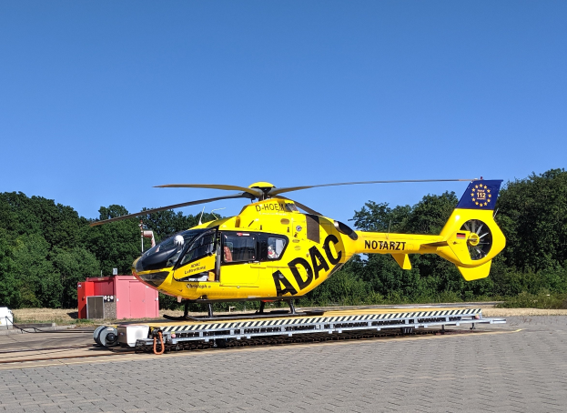 Gelber Hubschrauber auf einem Flughafenvorfeld mit Bäumen und einem klaren blauen Himmel, umgeben von Gegenständen, die wahrscheinlich mit Luftrettungsdiensten in Deutschland zu tun haben.