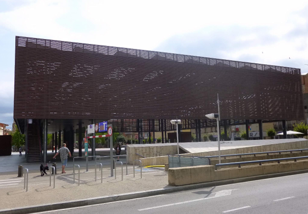 Großes modernes Gebäude mit vielen Fenstern als neuer Eingang zur Stadtmitte, umgeben von Straßeninfrastruktur, Fahrzeugen, Fußgängern, Bäumen und einem bewölkten Himmel.