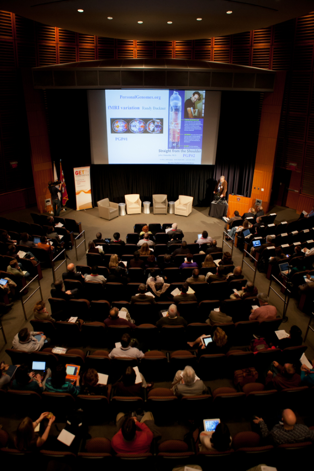 Ein mit Sitzenden gefüllter Saal, der Gegenstände hält, beleuchtet von vielen Lichtern, mit Fahnen auf der linken Seite, einem Podium auf der rechten Seite und einer Werbetafel auf der linken Seite.