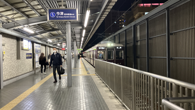 Ein Zug fährt auf einem Bahnsteig ein, mit Menschen in der Nähe, einem Geländer auf der rechten Seite, Gebäuden im Hintergrund, Bahnsteigbeleuchtung und einer Anzeigetafel mit Text.