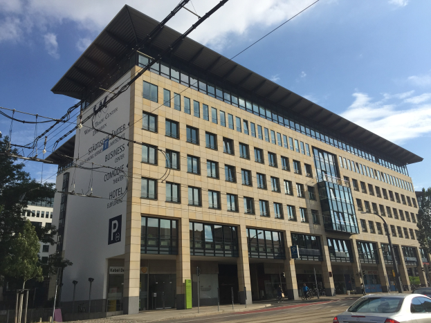 Großes modernes Gebäude mit zahlreichen Fenstern, beschriftet mit "Stadthalle Center für Geschäft", umgeben von Bäumen und städtischer Infrastruktur, mit Menschen und Fahrzeugen auf der Straße.