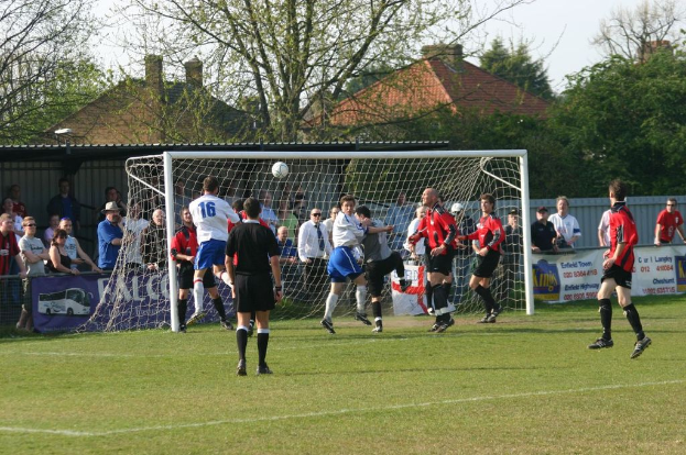 Spieler spielen Fußball auf einem Feld mit einem Tor hinter ihnen, während Zuschauer zuschauen; Bäume und Häuser sind im Hintergrund sichtbar.