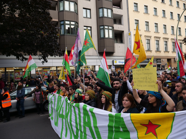 Große Gruppe von Menschen marschiert auf einer Straße mit Fahnen und Schildern, mit einem geparkten Auto auf der rechten Seite und einem Baum auf der linken Seite, vor Gebäuden mit Fenstern und Schildern, was auf eine algerische Demonstration hinweist.