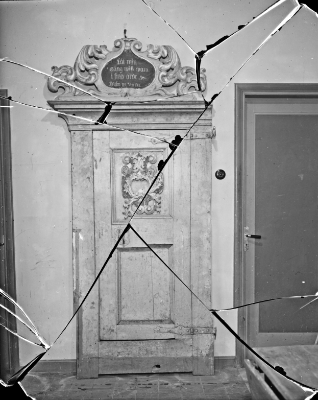 Ein Schwarz-Weiß-Foto einer zerbrochenen Tür mit verstreuten Glasscherben in einem Raum mit einer Wand im Hintergrund.