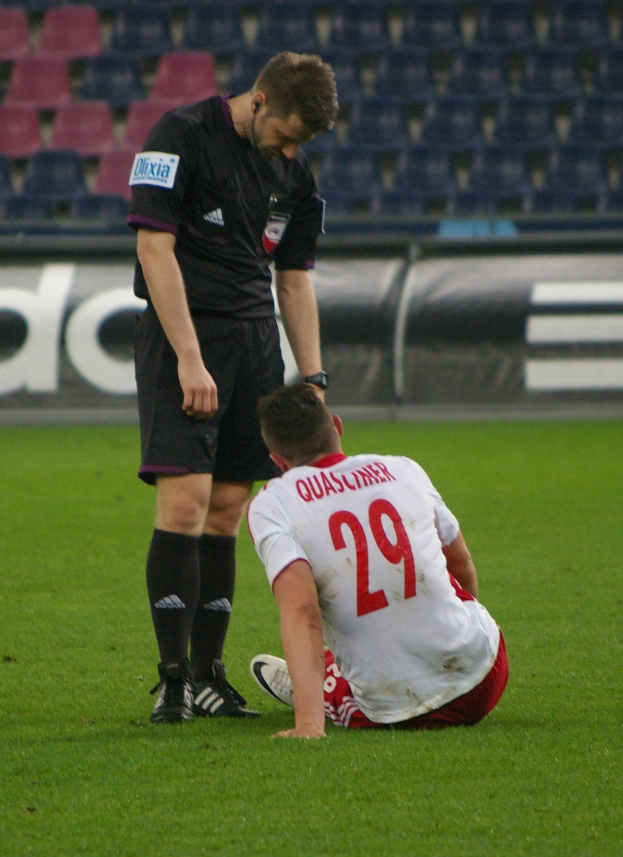 Ein Fußballspieler sitzt neben einem Schiedsrichter auf dem Boden in einem Stadion, beide in Sportkleidung, mit Schildern und Stühlen im Hintergrund.