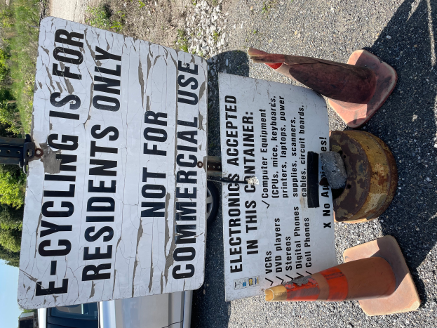 Ein Schild mit der Aufschrift "E-Bikefahren nur für Anwohner" steht an der Seite einer Straße, umgeben von Verkehrskegeln und anderen Gegenständen, mit einem Fahrzeug, Bäumen und einem klaren blauen Himmel im Hintergrund.