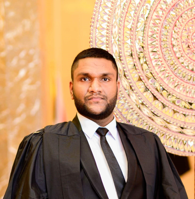 Ein Mann in schwarzer Robe, weißem Hemd und schwarzer Krawatte steht vor einer goldenen und cremefarbenen Wand, dargestellt als "Der Richter des Jahres".