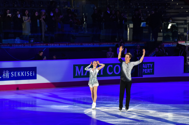 Zwei Personen beim Eislaufen auf einer Eisfläche mit Zuschauern und Fotografen im Hintergrund bei künstlicher Beleuchtung.