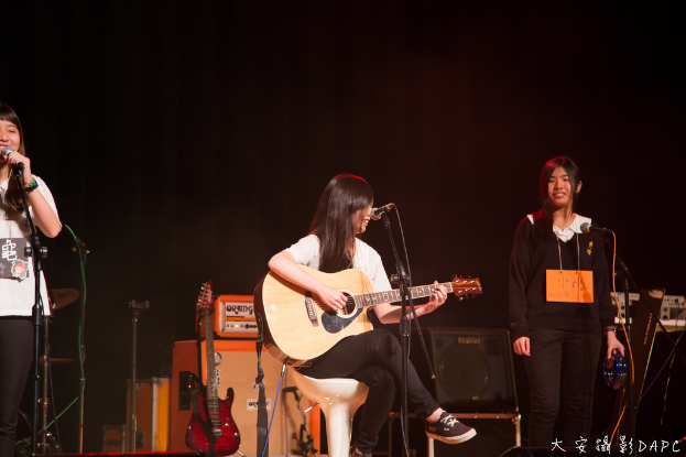 Drei Mädchen auf der Bühne, zwei singen in Mikrofone und eines hält ein Musikinstrument, mit einer Gitarre, einem Lautsprecher und einem Schaltpult.