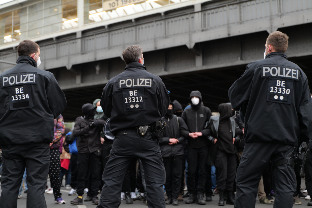 Eine Gruppe uniformierter Polizisten steht vor einer Menge von Menschen in schwarzen Uniformen und Masken mit einer Brücke und einem Gebäude im Hintergrund während einer Demonstration in einer Stadt.