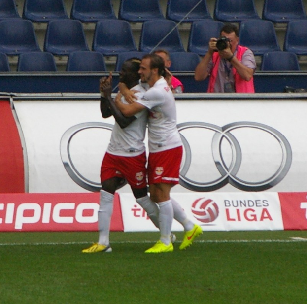 Zwei Fußballspieler feiern ein Tor auf einem Feld mit Zuschauern, einem Fotografen, einer Fahne, Stühlen und einer Wand im Hintergrund.