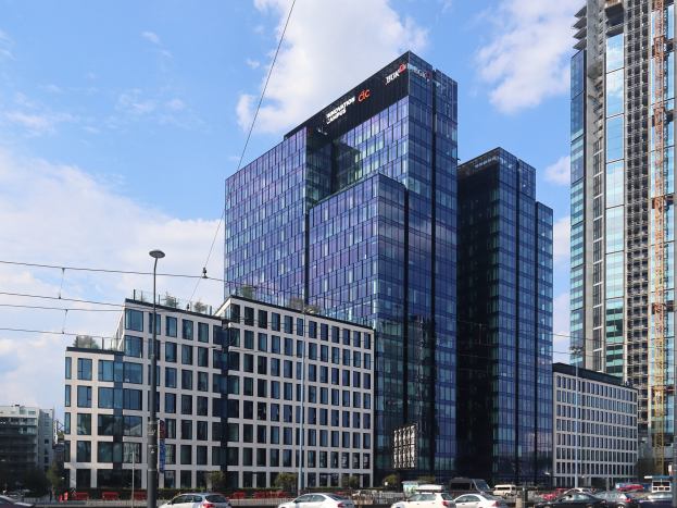 Hauptsitz der Deutschen Bank in Frankfurt, Deutschland, ein großes mehrstöckiges Gebäude mit zahlreichen Fenstern, umgeben von städtischer Infrastruktur wie Straßenlaternen, elektrischen Kabeln, Fahrzeugen, Grünflächen und einem bewölkten Himmel.