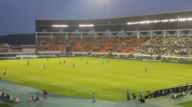 Ein Fußballspiel wird in einem Stadion mit Zuschauern auf Sitzen und Stehplätzen, einem Tor in der Mitte, einem Hügel und einem klaren blauen Himmel im Hintergrund und hellen Stadionlichtern gespielt.