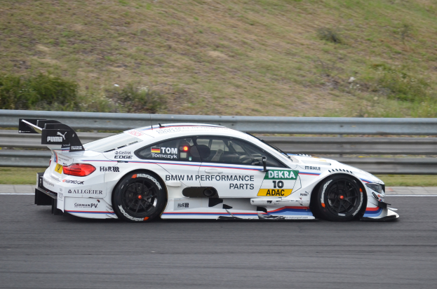 Ein BMW M4 DTM-Rennwagen auf einer Rennstrecke fahrend, mit Text auf dem Auto und einem Geländer im Hintergrund, umgeben von saftigem Grün.