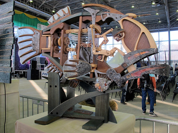 Große Metallfischskulptur in einem Innenraum mit Menschen, Stühlen, einem Zaun, einer Tafel, einem Lautsprecher, Vorhängen und Deckenlampen.