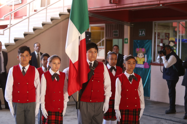 Eine Gruppe von Kindern in Schuluniformen steht zusammen, einer hält eine Fahne, mit Menschen, einer Kamera, einem Gebäude, einem Schild, einer Fahne und einer Treppe mit Geländer im Hintergrund.