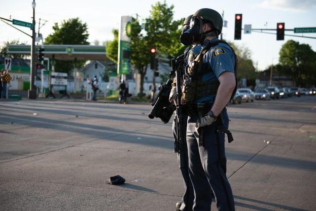 Polizeibeamter in voller Einsatzausrüstung mit einer Waffe an einer Straßenkreuzung mit Fahrzeugen, Verkehrszeichen, Schildern, Bäumen, Gebäuden und einer Tankstelle im Hintergrund.
