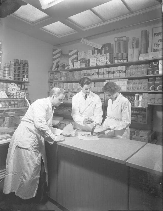 Drei Männer in weißen Kitteln an einer Apotheken-Theke mit Papieren und Gegenständen, auf einem Schwarz-Weiß-Bild.