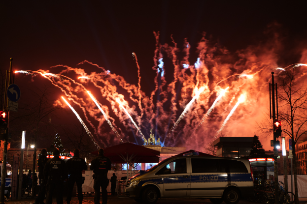Ein Polizeiwagen steht vor einer Menschenmenge, mit beleuchteten Gebäuden und Bäumen im Hintergrund, während farbige Feuerwerke den Himmel erhellen, während die Silvesterfeierlichkeiten in Berlin stattfinden.
