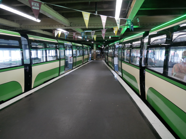 Grüner und weißer Zug mit Fahrgästen an einer Bahnstation in München vorbei, Decke mit Lampen und dekorativen Fahnen.