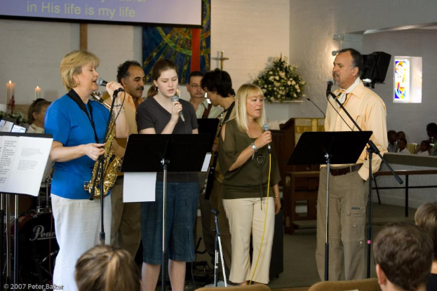 Eine Gruppe von Menschen versammelte sich in einem Raum mit einer Wandbanner, Kerzen, Papier, Mikrofonen, einem Foto Rahmen und Blumen.