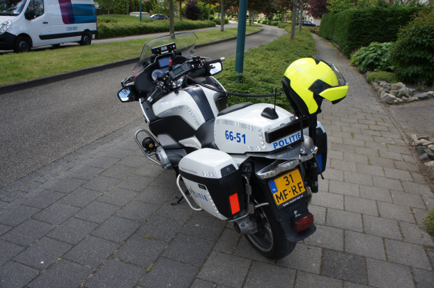 Ein Polizei-Motorrad am Straßenrand mit einem Helm darauf, umgeben von Bäumen, Gebäuden und anderen Fahrzeugen.