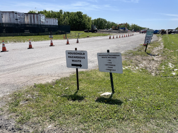 Straßenrand-Sammelstelle für "Haushaltsgefährliche Abfälle" markiert durch Verkehrskegel, mit Fahrzeugen, Gras, Bäumen und bewölktem Himmel im Hintergrund.