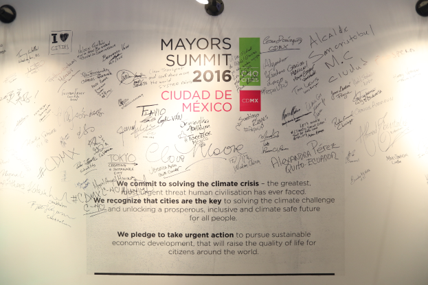 Eine weiße Wand mit einem Schild, auf dem "MAYORS SUMMIT 2016" steht, beleuchtet von Deckenlampen.
