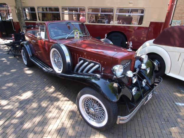 Ein Oldtimer neben einem Doppeldeckerbus auf einem Kopfsteinpflasterweg geparkt, mit Stühlen und einem Baum im Vordergrund und einem Gebäude mit Fenstern im Hintergrund; Menschen sind im Bus zu sehen.